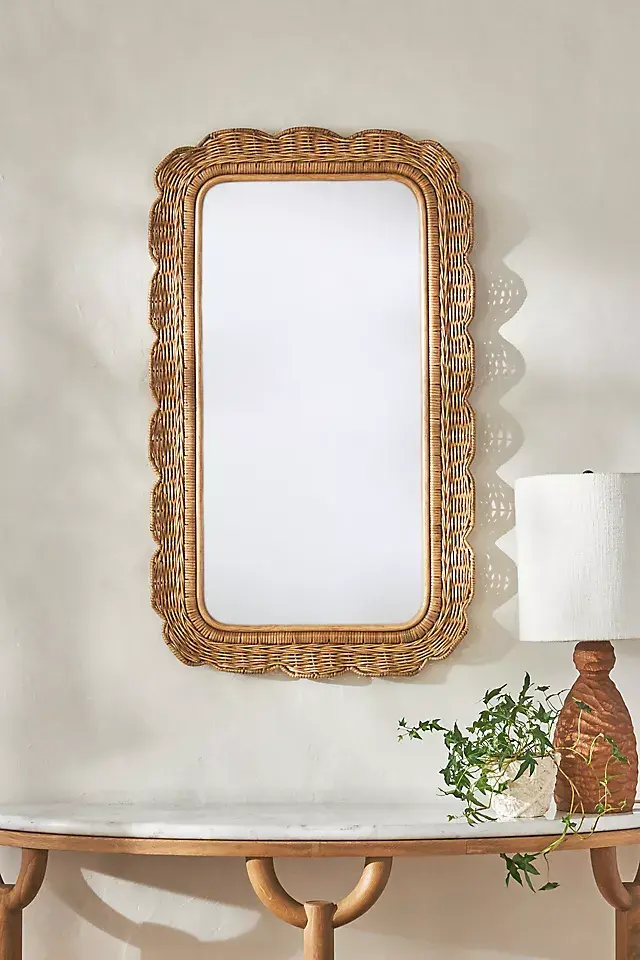 Wicker-framed mirror on a cream wall above a wooden table, with a lamp and leafy plant beside it in a calm, airy room.