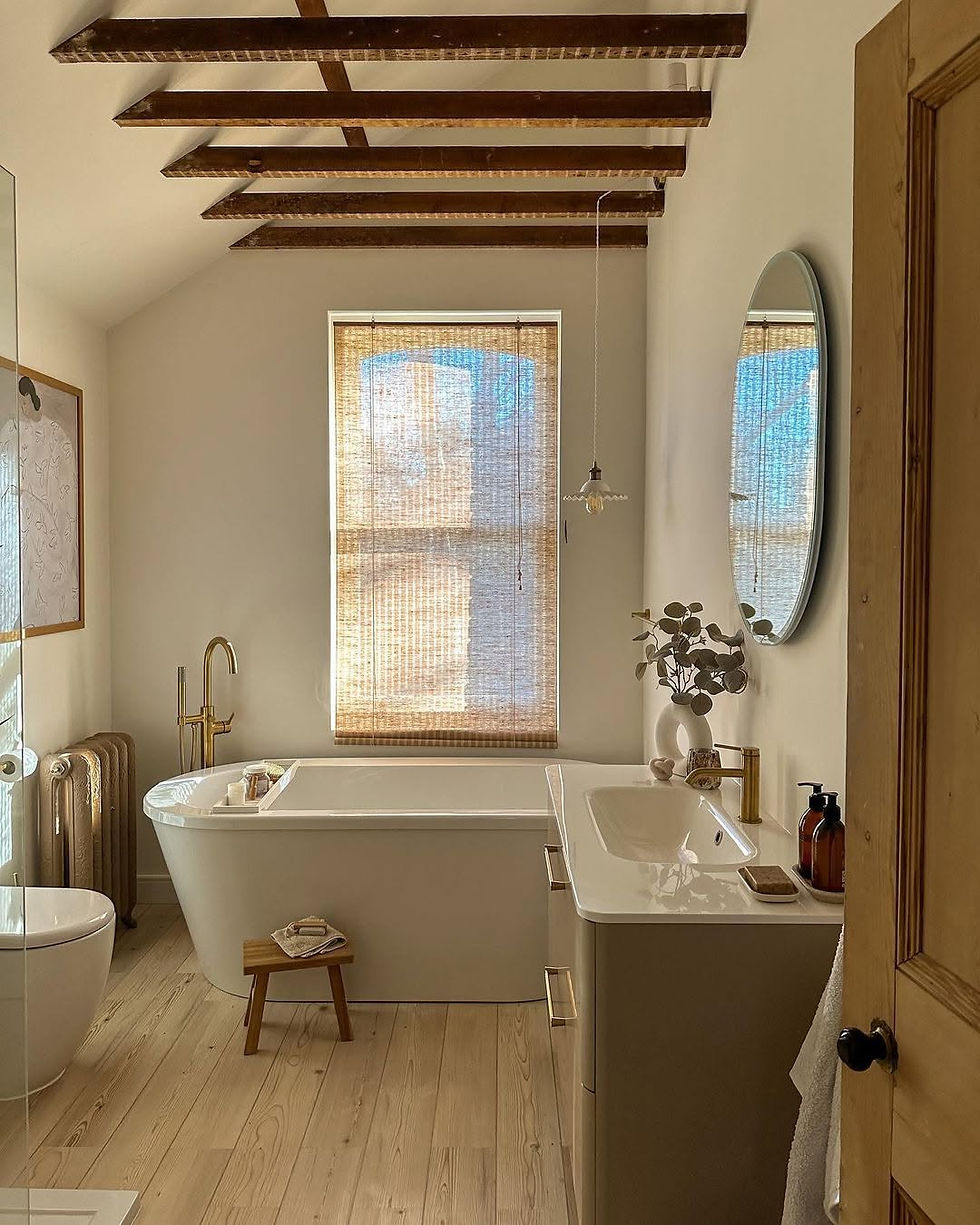 Warm minimalist bathroom with freestanding tub, brass fixtures, vanity and oval mirror, sunlit through woven blinds.