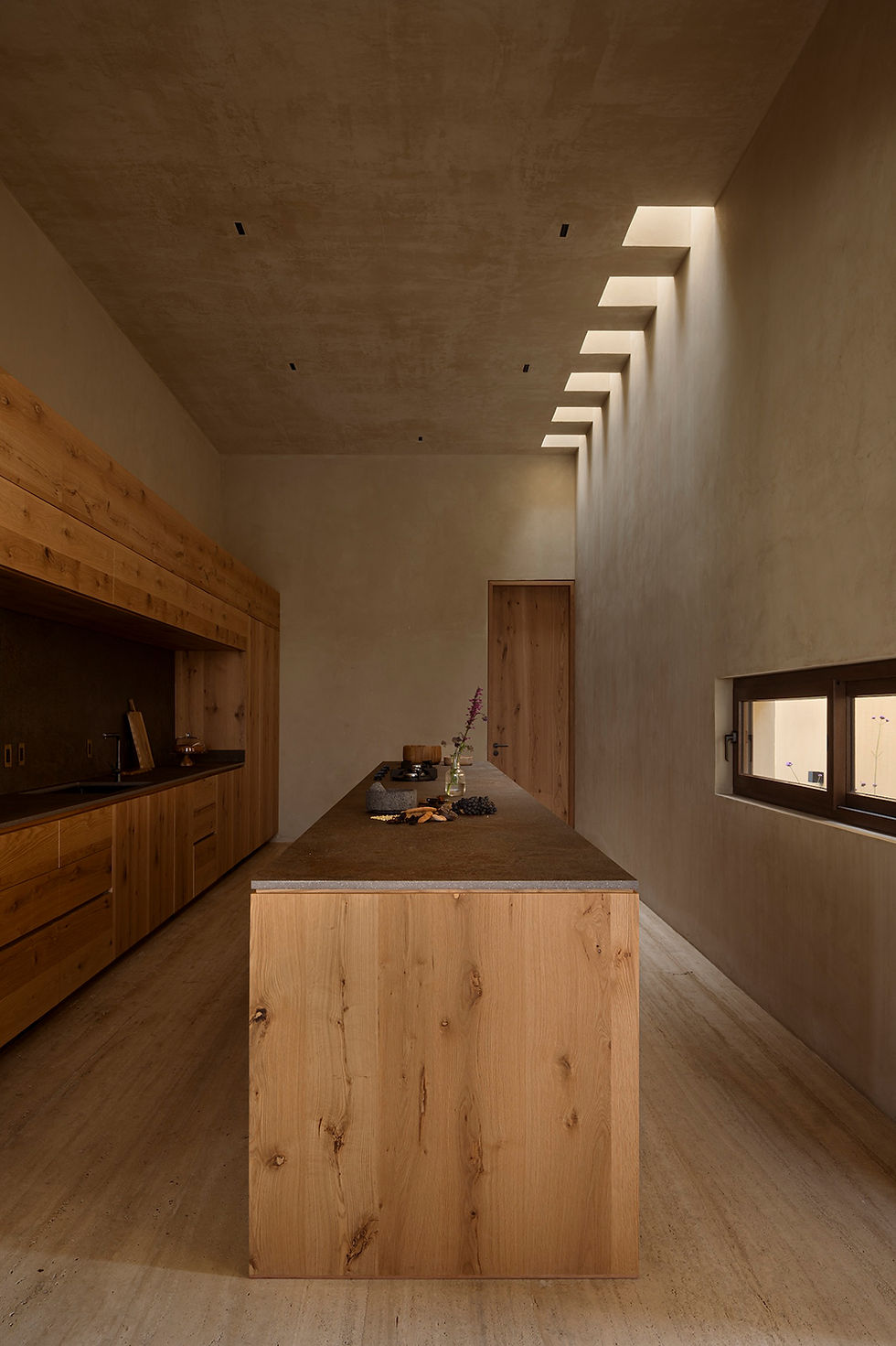 Minimal wood kitchen with island, beige plaster walls, skylight openings, and warm natural light; no people present