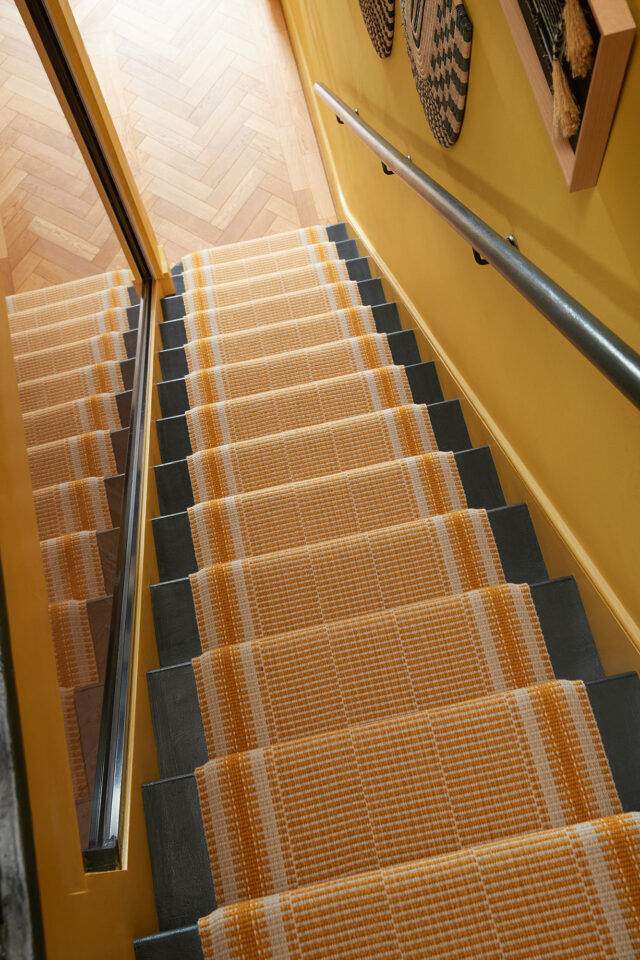 Top view of a yellow staircase with orange striped carpet runner, black handrail, and herringbone wood floor.
