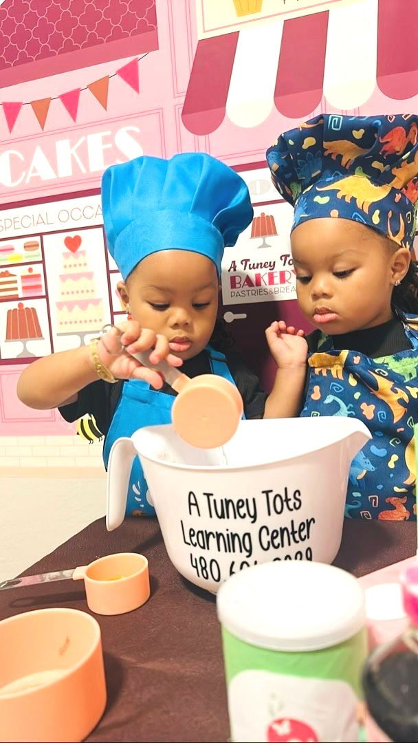 2 kids wearing a black shirt, wearing a chef-printed blue hat, playing a chef. There are baking materials and ingredients in front of them, and one of the kids is holding a cup, pouring water on a mixing bowl