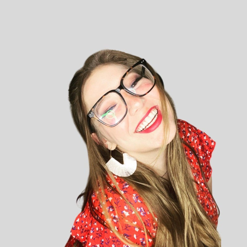 Smiling woman with glasses and red floral dress against gray background. Wearing large white earrings, exuding joy and warmth.