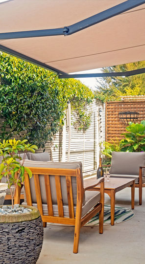 A courtyard garden with some outdoor chairs and a low table. Surrounding the furniture are various pots and planters with lush green plants. Shading the area is an outstretched awning.