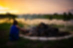 A little girl roasts marshmallows by a campfire and a pond with a sunset in the background.
