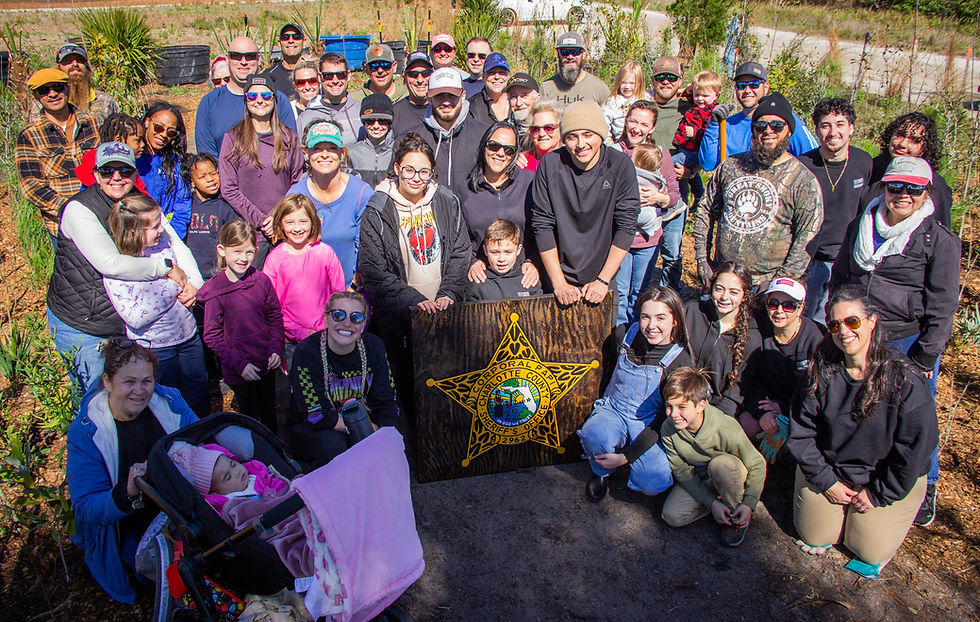 A diverse group of people posing outdoors around a large wooden badge reading "Charlotte County Sheriff's Office." Bright, sunny day.