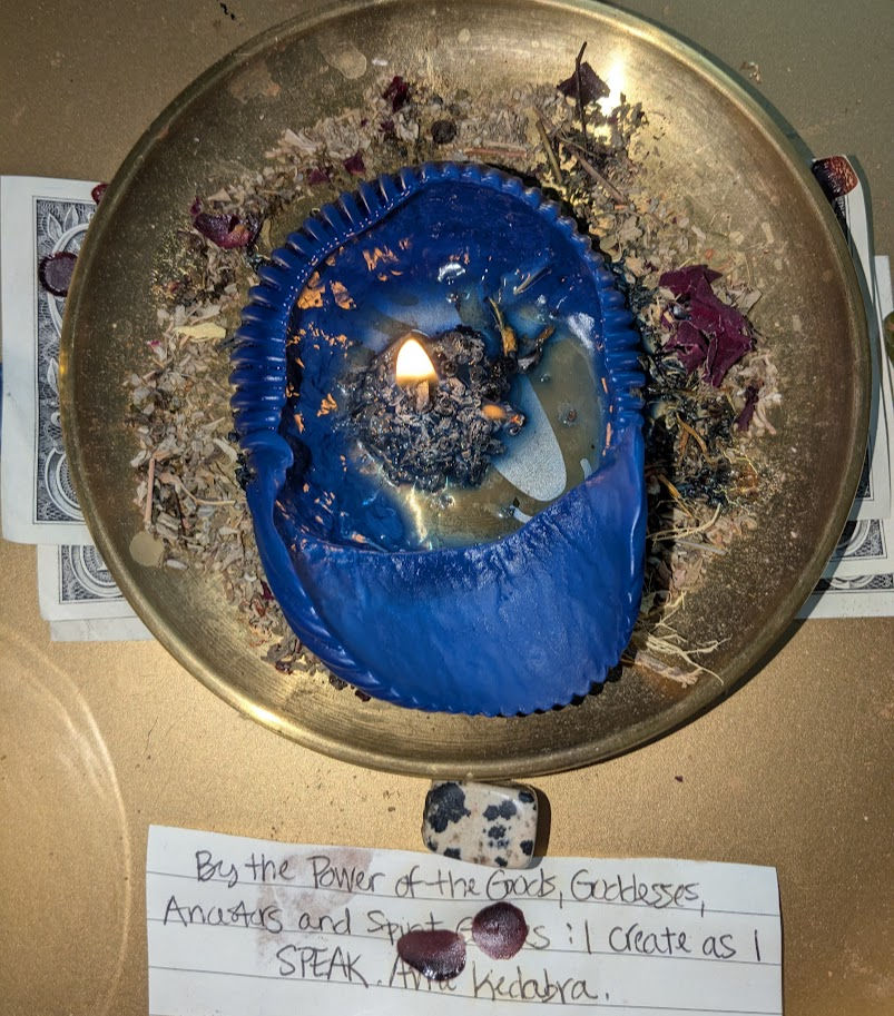 Blue candle melting on a plate with herbs and petals, surrounded by dollar bills. A handwritten note reads: By the Power of the Gods.