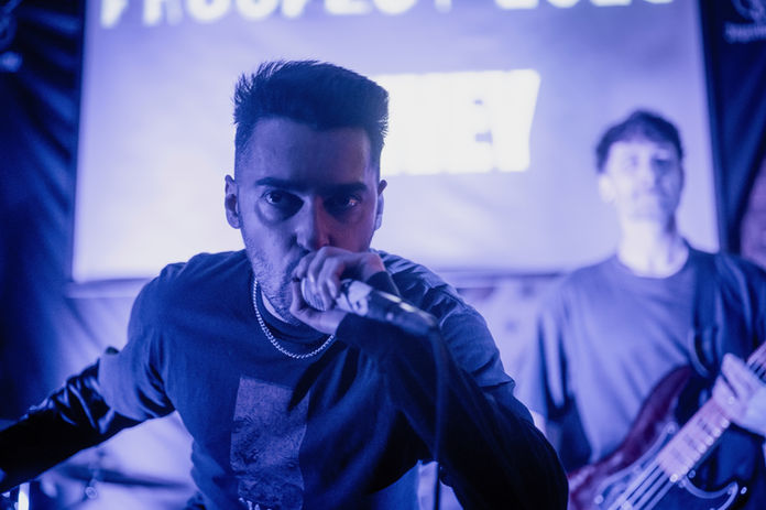 A close up face of a singer in Convey live on stage, with him holding his microphone and leaning forward to the camera with a guitarist in the background