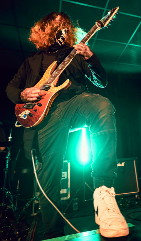 A photo of a guitarist with his foot close to the microphone and him playing the guitar, with a mask on.