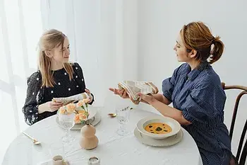 Two women sit at a round table with a white tablecloth, exchanging plates. One holds a book. Soft lighting, with flowers and soup on the table.