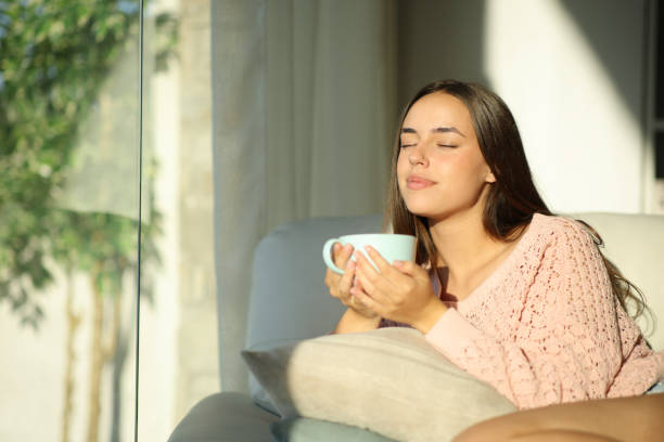 Woman in a pink sweater enjoys a cup of tea, sitting on a couch by a window with sunlight streaming in. Relaxed and content mood.