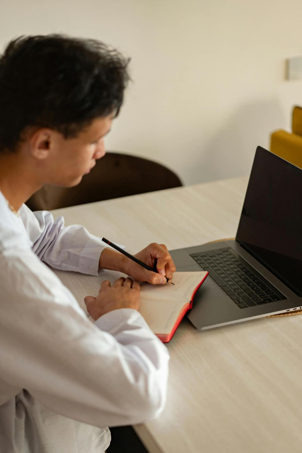 A person writing in a notebook next to a laptop on a light-colored table. The setting is minimal with a focus on concentration.