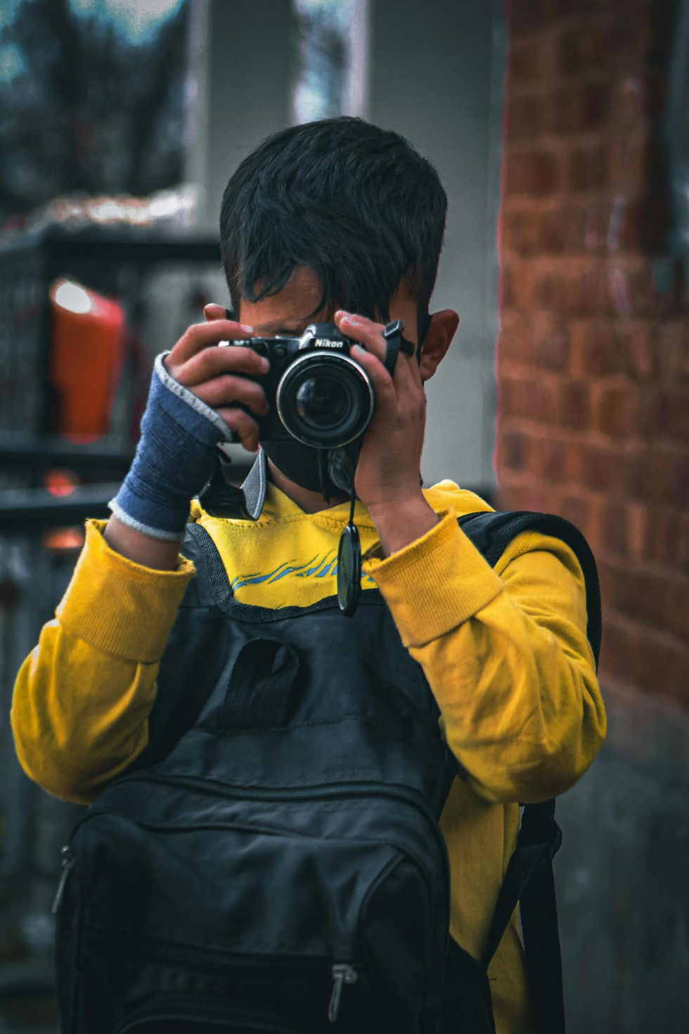 Young boy in a yellow sweater and backpack takes a photo with a camera. He has a blue bandage on his left hand. Brick wall in background.