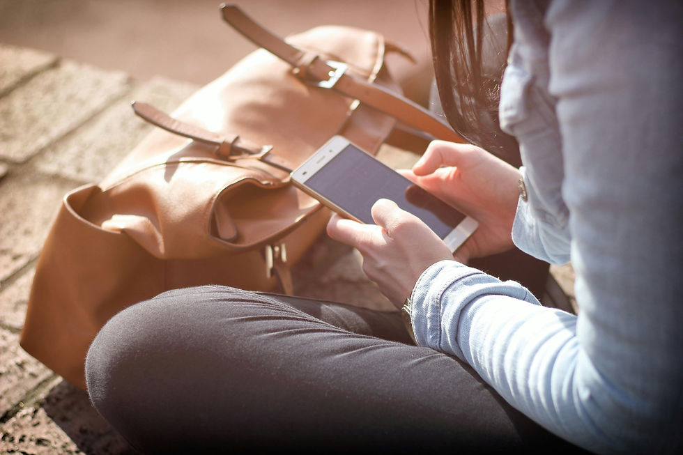 Person in jeans using a smartphone, seated outdoors next to a brown leather bag on steps. Warm sunlight creates a relaxed mood.