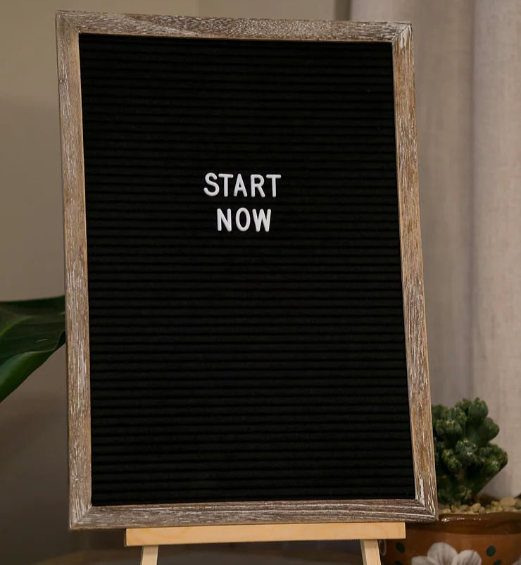 Wood-framed black letter board displaying "START NOW" in white text, set against a neutral background with a potted plant nearby.