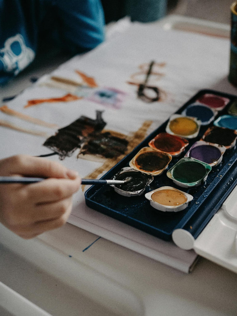 Hand painting with watercolors on paper, vibrant colors, and brushes visible. Casual setting with relaxed, creative mood.