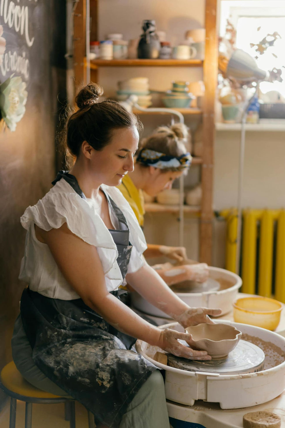 Two women sculpt pottery at a wheel in a sunlit studio. Shelves hold ceramics. The woman in front wears a white blouse; focused and calm.