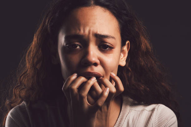 A woman with curly hair looks distressed and tearful, hands near her face, against a dark background. She wears a light ribbed sweater.