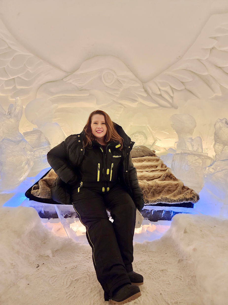 Woman smiling, sitting on a fur-lined ice bench in an ice room. Large bird carving on the wall. Cozy and bright atmosphere.