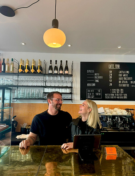 Die Eigentümer Sinah und Matthias lächelnd hinter der goldenen Theke ihres Cafés.