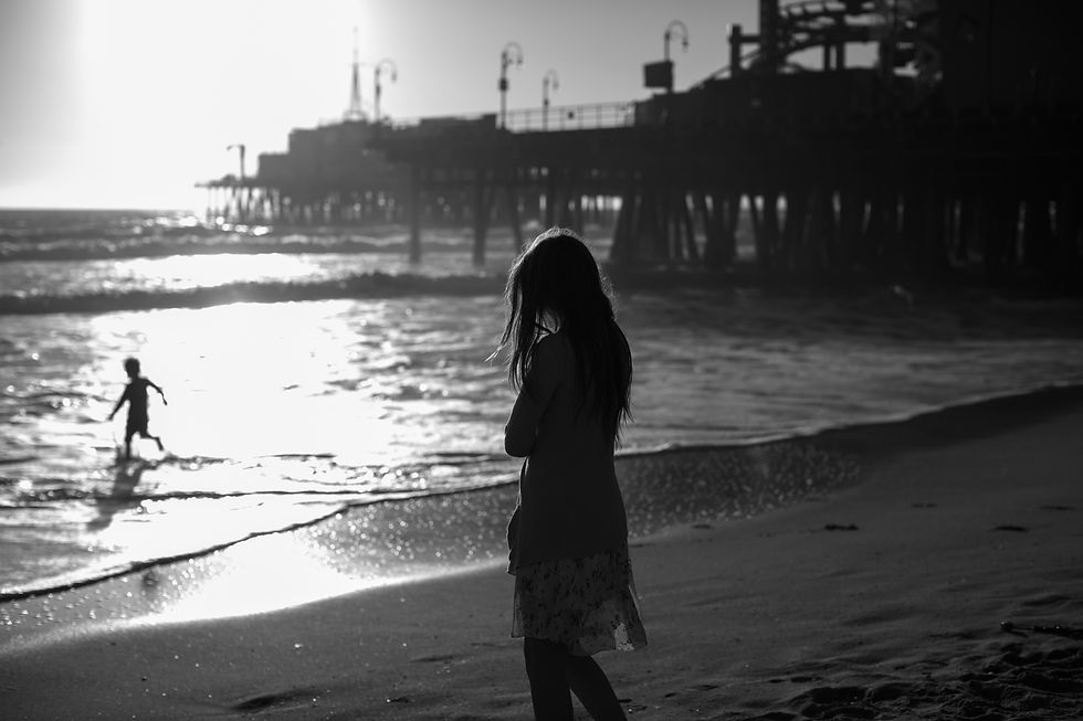 Silhouettes of a girl on the beach and a child playing in the waves, with a pier in the background at sunset. The mood is serene.
