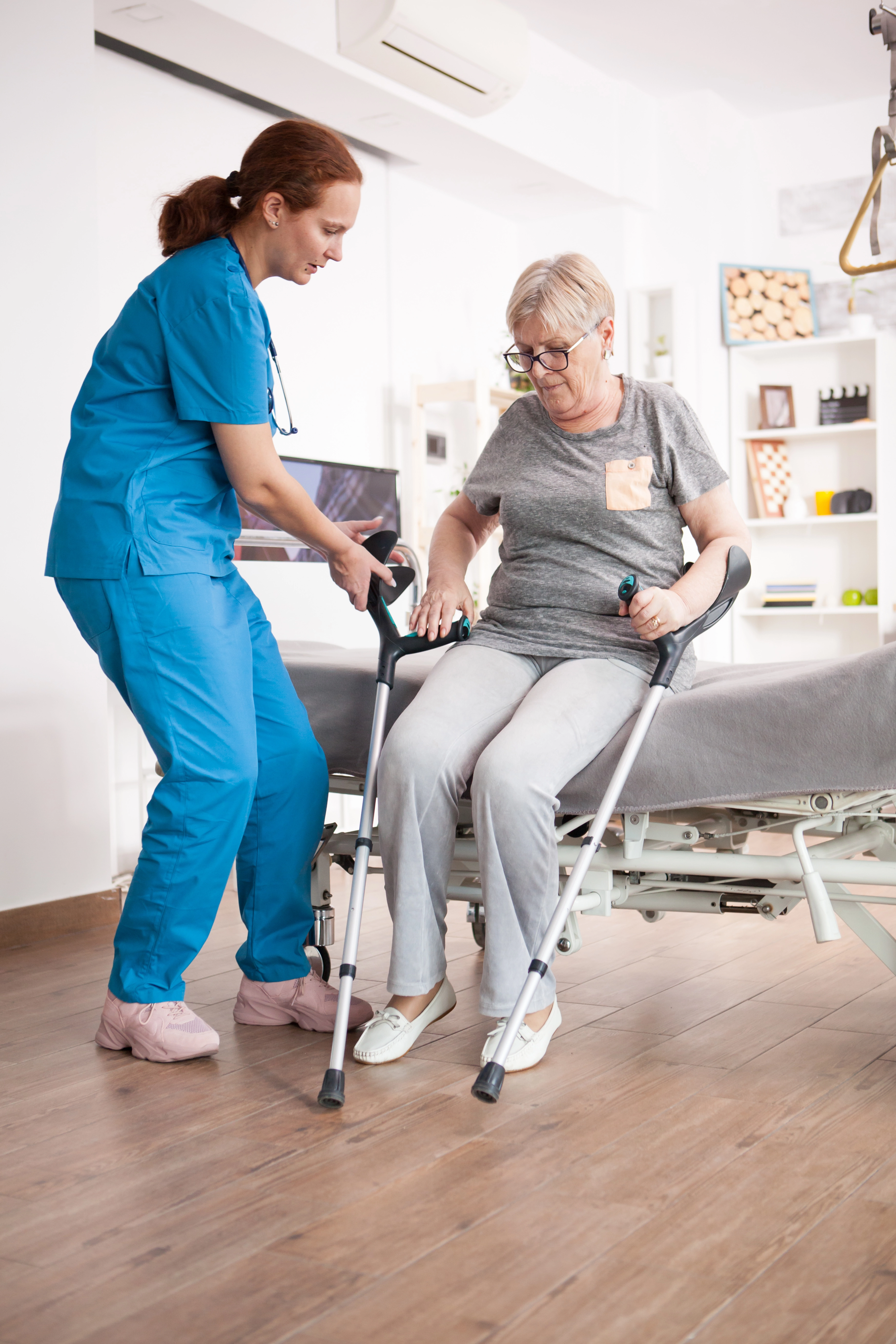 nurse-helping-old-woman-sit-bed-nursing-home-after-walking-with-crutches.webp