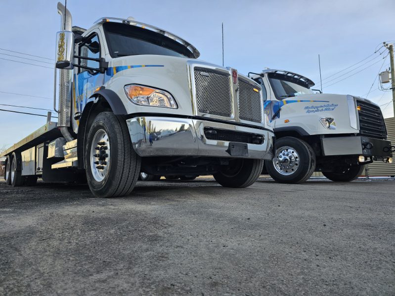 Deux camions de remorquage vus de face, avec cabines blanches et détails bleus, stationnés sur une surface asphaltée.