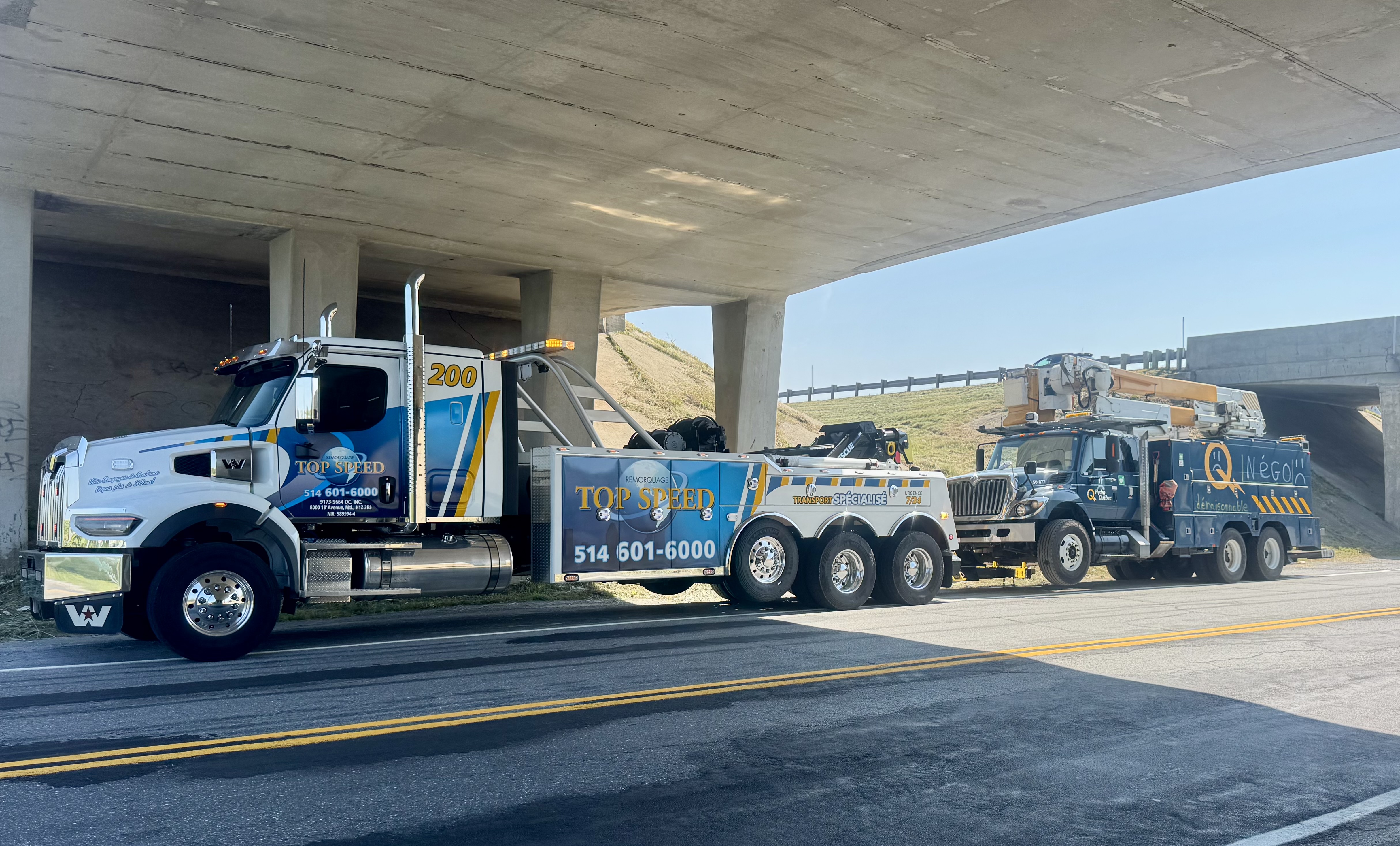 Dépanneuse lourde remorquant un camion à nacelle sous un viaduc en béton.