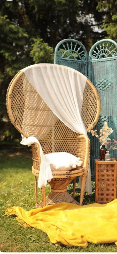 A decorative outdoor seating setup with a large wicker peacock chair draped in white fabric, a yellow blanket spread on the grass, a small side table with dried flowers in amber bottles, and a blue-green wicker backdrop, all surrounded by greenery.