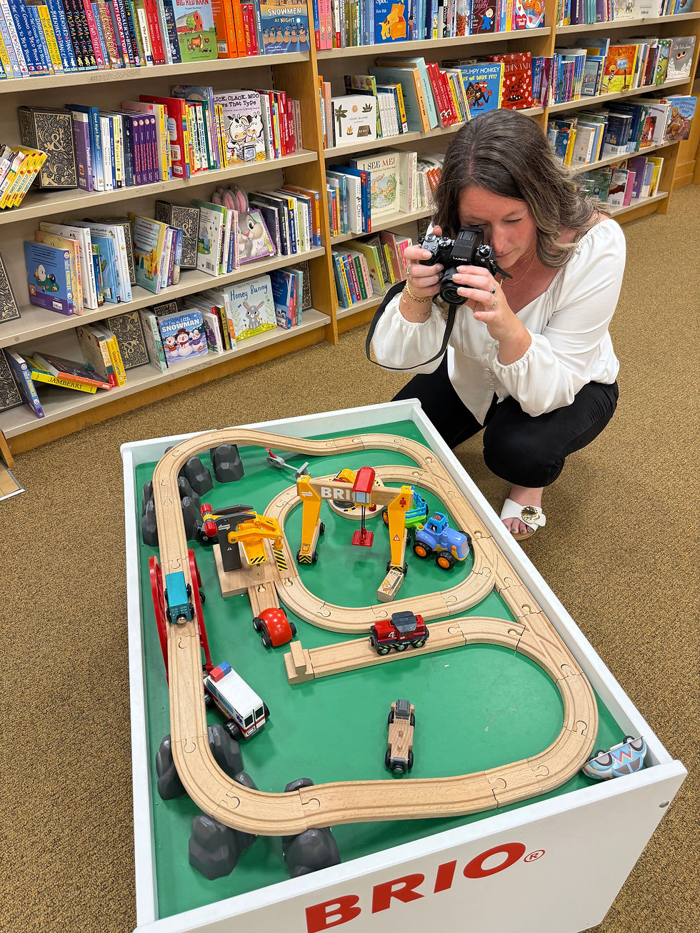 Woman photographing a BRIO wooden train table in a children's bookstore aisle filled with colorful picture books and educational titles
