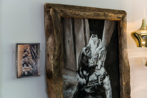 Bild mit Lehmfarben gemalen und mit Holzrahmen. Darauf wird ein Hund unter der Schnauze von einer Hand gestreichelt.