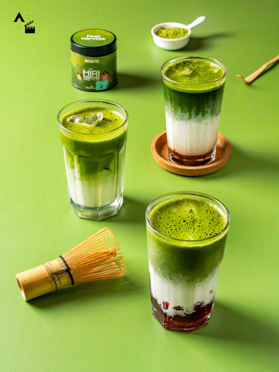 High-angle commercial shot of three different iced matcha latte variations in clear glasses, styled alongside a bamboo whisk on a solid green studio backdrop.
