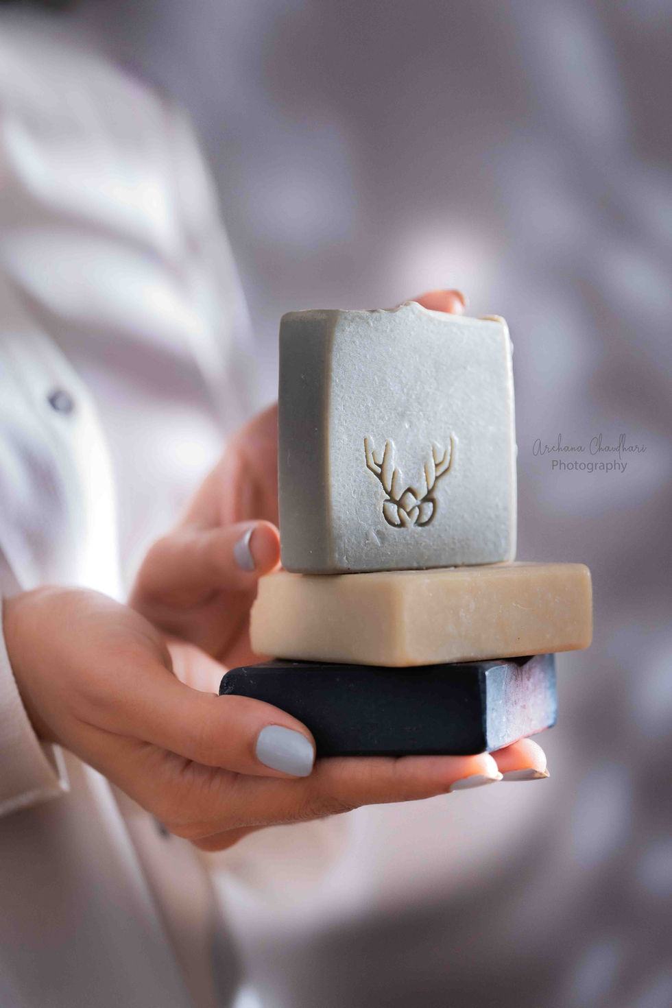 A side view of a hand holding a vertical stack of three pastel and charcoal artisanal soap bars under dappled leaf shadows and soft focus sunlight.