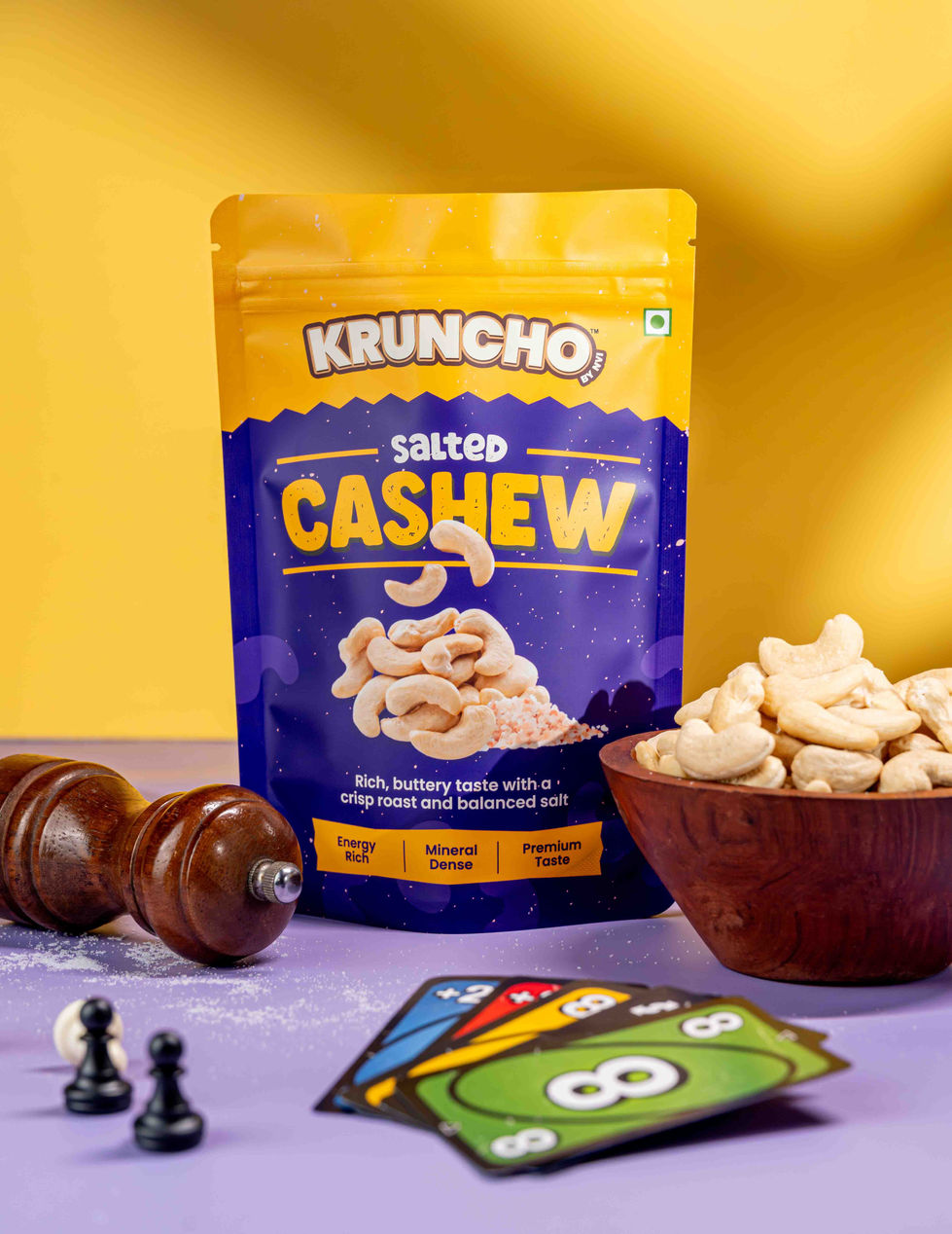 Creative product shot of Kruncho Salted Cashew pouch against a contrasting bright yellow background, styled with a wooden bowl, pepper mill, and Uno cards.