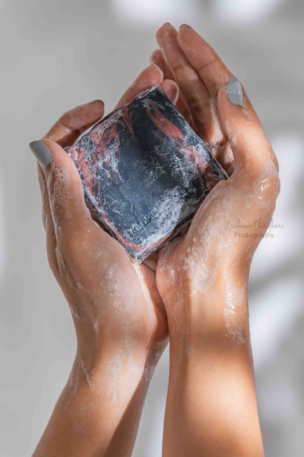 Wet, soapy hands cupping an engraved black activated charcoal bar soap with rich red clay swirls, covered in fine white lather bubbles against a clean backdrop.