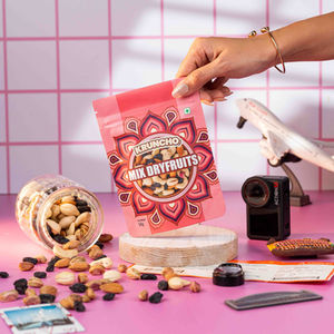 Commercial box packshot of Kruncho Mix Dryfruits displayed in an open pink display box, styled with scattered nuts, a mini airplane, and glass jar props by Acube Studios.