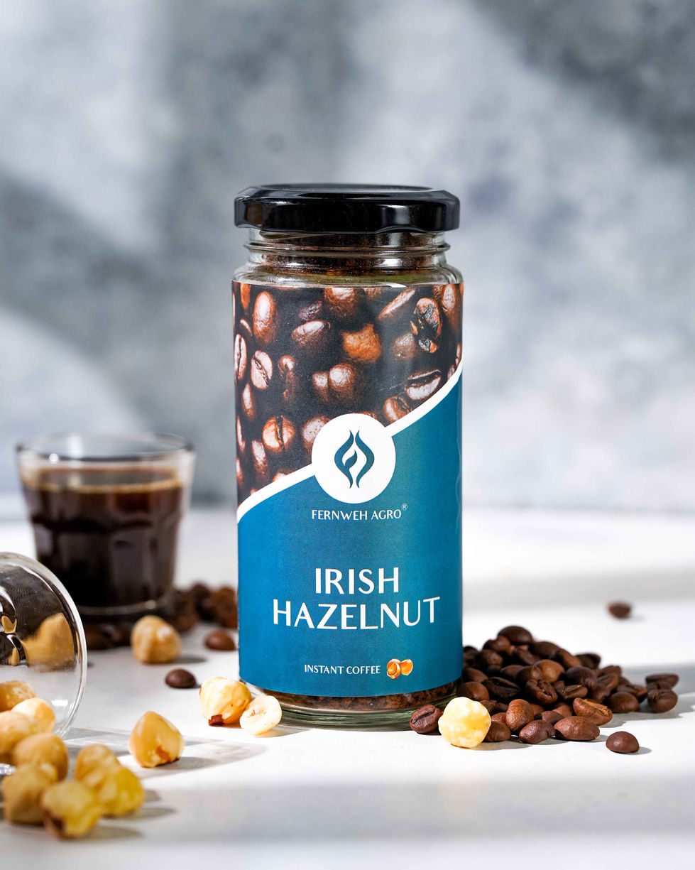 A sharp product shot showcasing the Fernweh Agro Irish Hazelnut instant coffee bottle positioned alongside peeled whole hazelnuts, scattered coffee beans, an espresso shot glass, and a tall iced coffee milk drink on a bright table surface.