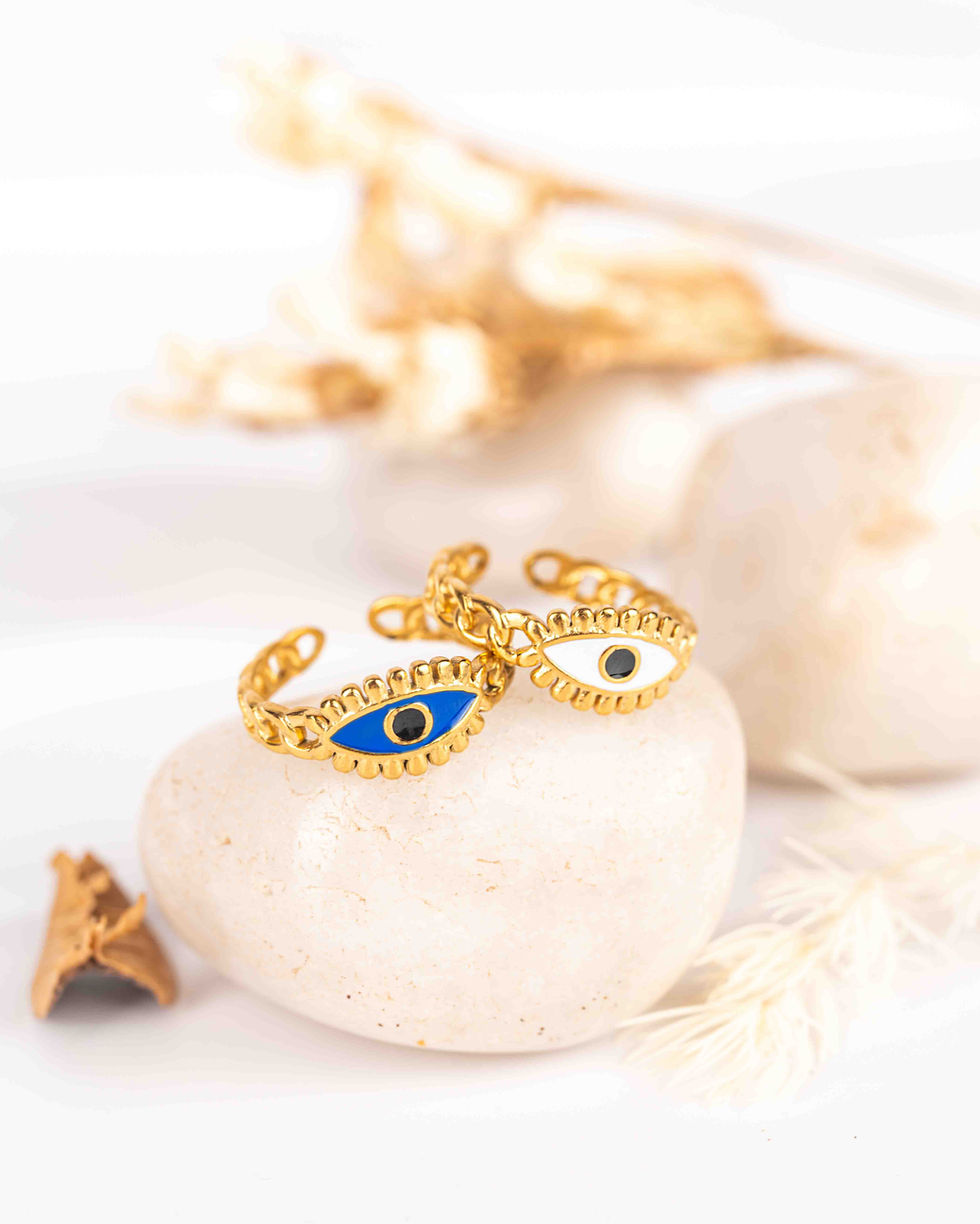 A macro close-up shot of two gold chain-link adjustable rings featuring blue and white enamel evil eye charms resting on a smooth white river stone.