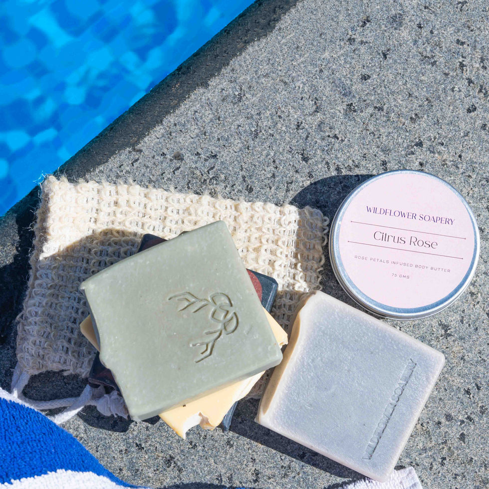 A high-angle product layout showing a sage green bar soap and a lavender bar soap arranged on a woven fiber pouch alongside a Citrus Rose Body Butter container by a pool edge.