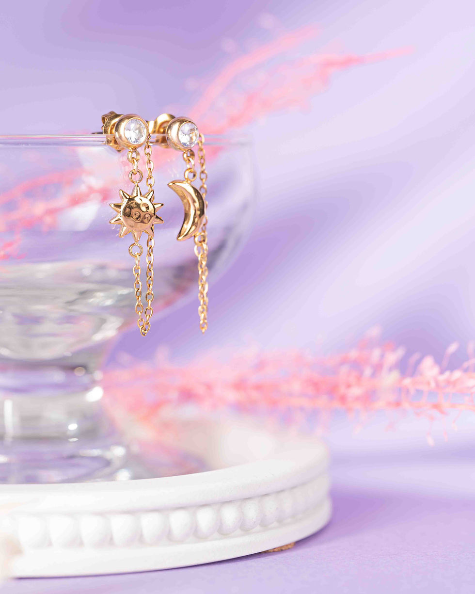 Gold celestial sun and moon dangle chain earrings hanging elegantly from the rim of a clear glass coupe on a soft lavender purple backdrop.