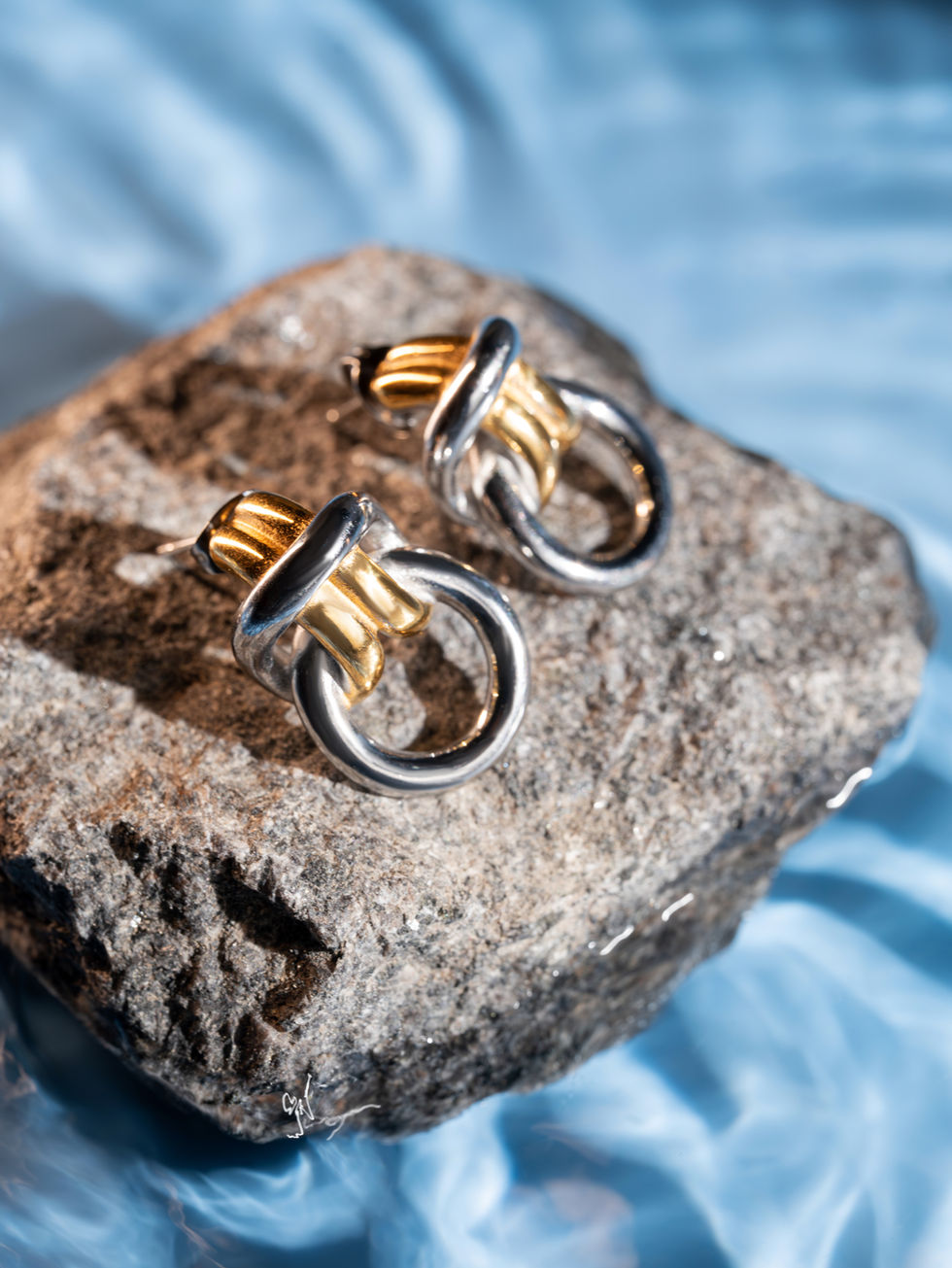 A pair of interlocking silver and gold chunky knot earrings resting on a textured gray rock submerged in shimmering, sunlit blue water ripples.