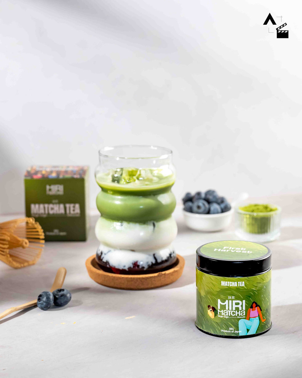 Crisp lifestyle product shot of an iced blueberry matcha drink next to a Miri Matcha tin, a bowl of fresh blueberries, and a bamboo chasen whisk on a gray stone texture.