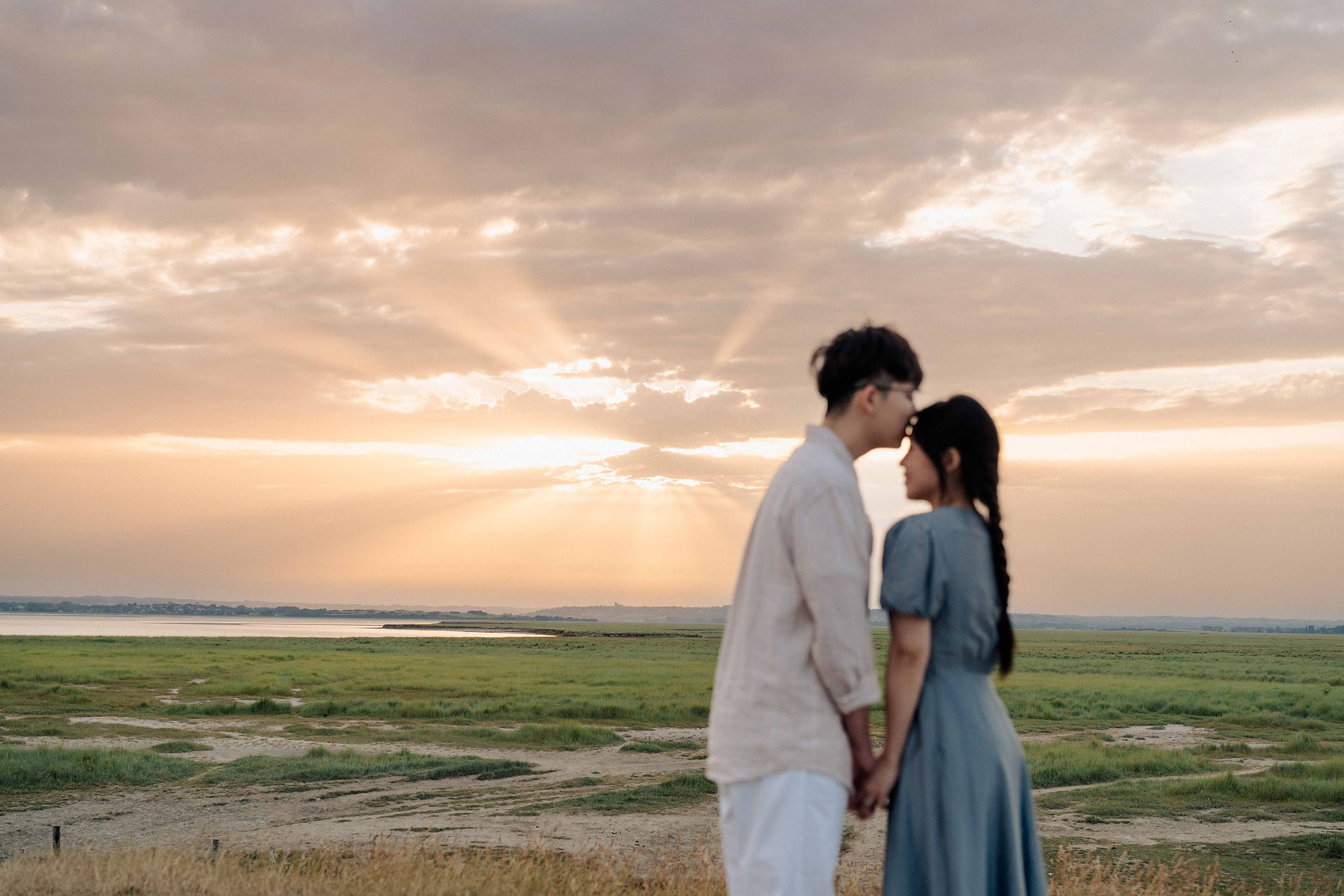 Man gently kissing his partner's forehead against a radiant golden sunset over the Normandy salt marshes