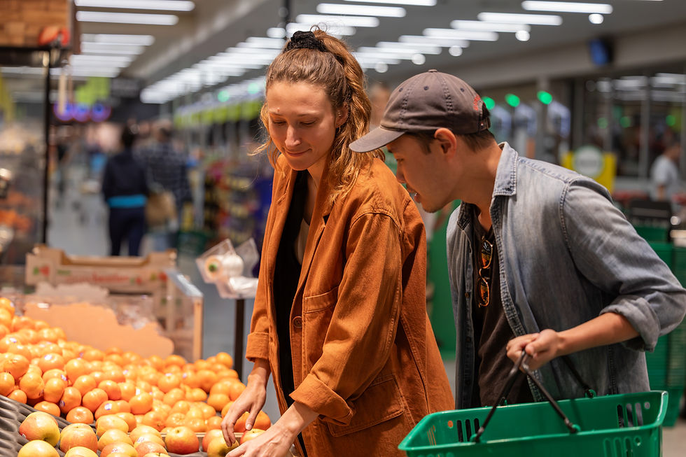 A woman in a brown jacket and a man in a cap browse apples in a grocery store. The background is busy with shoppers and bright lights.