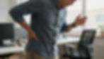 Middle-aged man holding his lower back and leaning forward in discomfort at the office, illustrating symptoms of low back pain when standing during daily activities.