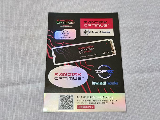 机の上に置かれた黒いSanDisk OPTIMUSの宣伝チラシ。SSD画像とDetonation FocusMeロゴ、TOKYO GAME SHOW 2026のQRコード付き案内文がある