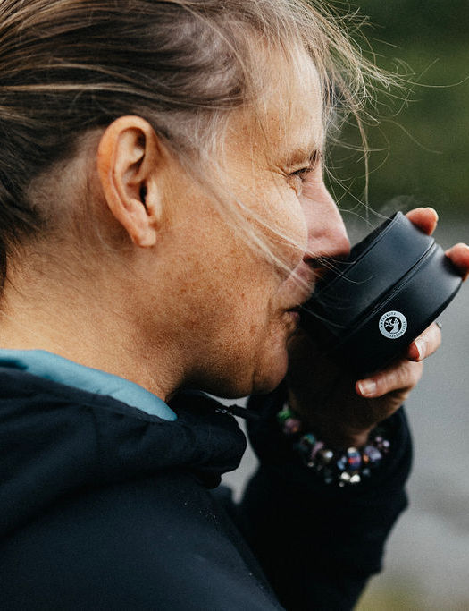Woman in outdoor clothing enjoying a warm drink from a wooden cup, with a blurred mountain lake in the background.