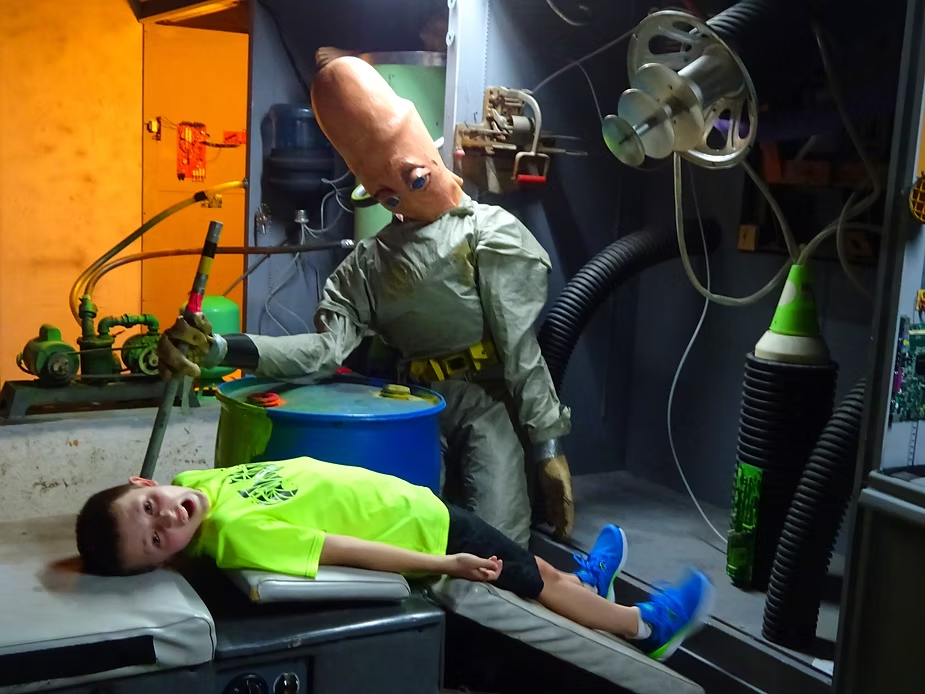 A child in a neon green shirt playfully lies on a table in a science fiction-themed room, with a large-headed alien mannequin beside him.