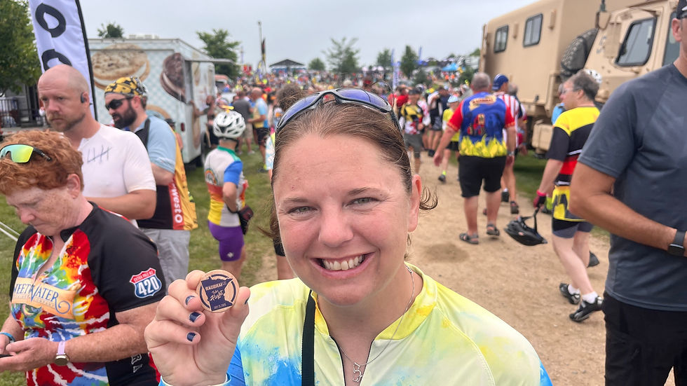 Smiling woman holds a small token. She's in a colorful cycling shirt at an outdoor event with many people in the background.