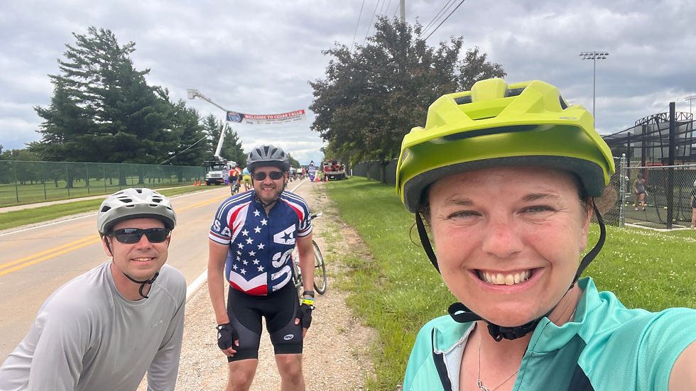 Three cyclists in helmets smile on a road near trees and a banner reading "Welcome to Cedar Falls." Overcast sky, cheerful mood.