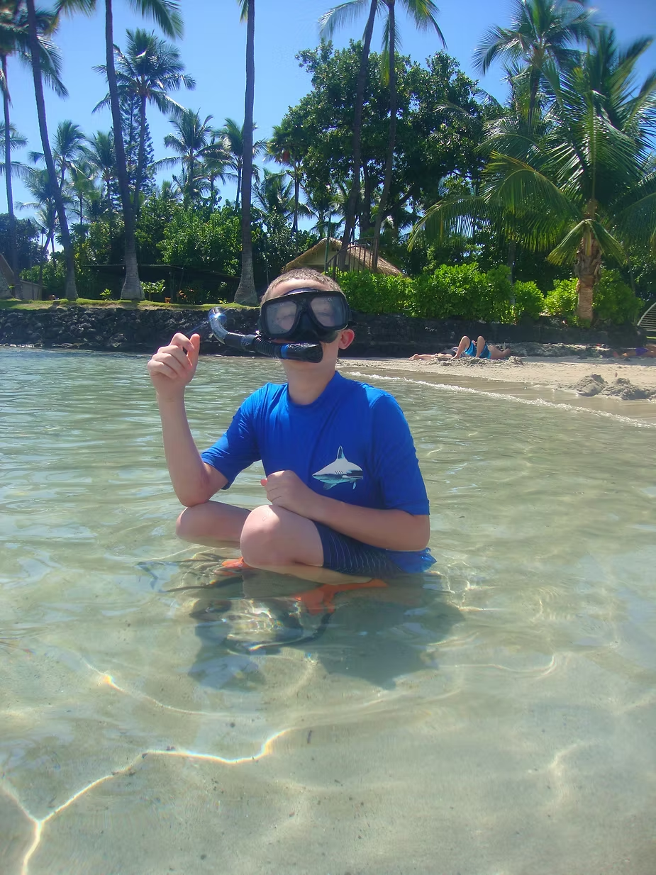 A person in a blue shirt and snorkel gear kneels in clear water on a sunny beach. Palm trees and sunbathers are in the background.